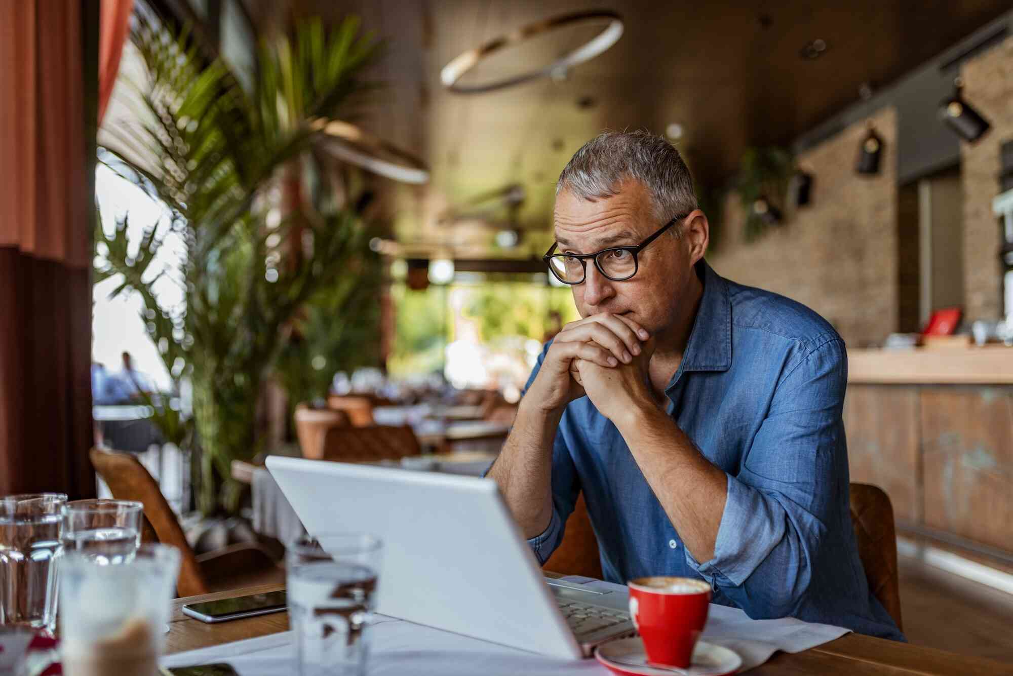 A man in a denim shirts sits at a cafe looking stressed while looking at his laptop.